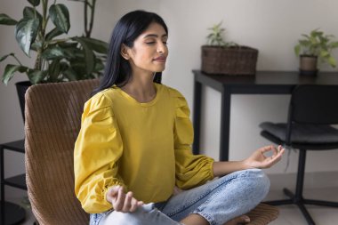 Sakin genç Hintli kadın evde meditasyon yapıyor, koltuğunda oturmuş meditasyon yapıyor, Zen Mudra 'yı elinde tutuyor, pratik yapıyor, barış yapıyor, konsantrasyon kuruyor, soğuk ve temiz havayı kapalı gözlerle soluyor.