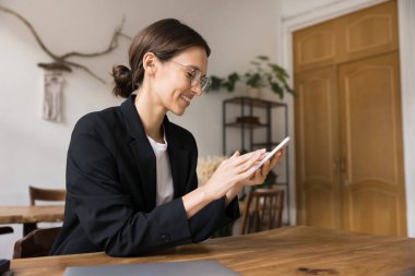 Resmi ceket giymiş güzel bir kadın gözlük takıyor. İş yerinde akıllı telefonuyla oturuyor, internet sohbetine öncülük ediyor. Şirket müşterilerine cevap veriyor.