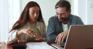 Positif orta yaşlı koca ve genç eş kağıt faturalarını, faturaları, faturaları evde birlikte kontrol ediyorlar, mali belgeleri gözden geçiriyorlar, masadaki harcamaları ve bütçeyi laptopla tartışıyorlar.