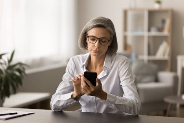 Gözlüklü, cep telefonuyla yazan, mesaj yazan, internet teknolojisini iş iletişimi için kullanan, akıllı telefon tutan, çalışma masasında oturan ciddi, olgun girişimci kadın.