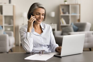 Mutlu orta yaşlı iş kadını cep telefonunda dizüstü bilgisayarla konuşuyor, cep telefonuyla konuşuyor, evden çalışmak için kablosuz teknolojiyi kullanıyor, gülümsüyor, sohbetten zevk alıyor.