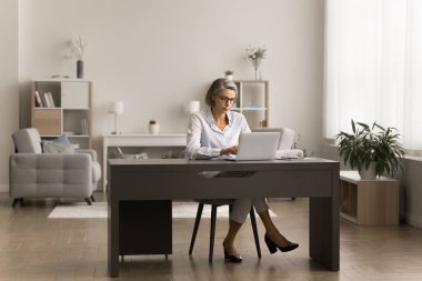 Ev bilgisayarında çalışan gözlüklü ciddi bir iş kadını oturma odasının iç tarafındaki masada oturuyor ve uzaktan kumanda için internet iletişimini kullanıyor.