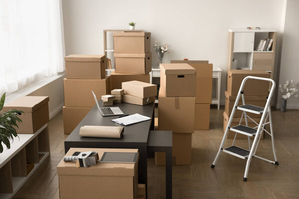 Рабочее место склада с ноутбуком на рабочем столе среди кучи картонных коробок и стремянки. Интерьер хранилища интернет-магазина с упакованными бумажными пакетами, никаких людей