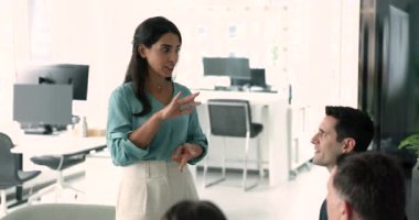 Mutlu Latin takım lideri kadın yöneticilerle toplantı yapıyor, meslektaşlarının önünde duruyor, bir grup işçiyle konuşuyor, görevleri tartışıyor, takım çalışması, strateji ve proje yönetimini planlıyor.