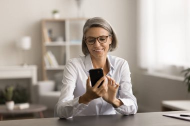 Gözlüklü mutlu iş kadını cep telefonuyla sohbet ediyor, akıllı telefonda mesaj yazıyor, online medya uygulaması kullanıyor, internette geziniyor, aletleri tutuyor, iş yerinde oturuyor.