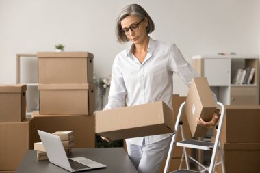 Olgun bir e-ticaret depolama işçisi elinde bir paket, çalışma masasında dizüstü bilgisayarla karton kutular taşıyor, nakliye alımları üzerinde çalışıyor, müşterilere paket gönderiyor.