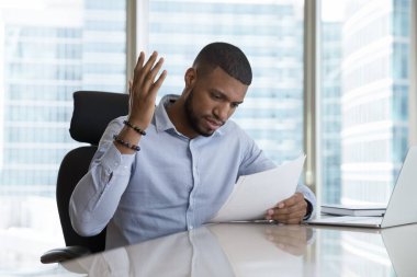 Frustrated African man read bad news in postal letter or paper document sit at desk in modern skyscraper office, get negative response, dismissal notice, high bills, huge taxes, bank debt notification clipart