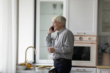 Yaşlı bekar adam akıllı telefondan konuşuyor, sağlıklı sebze hazırlıklarından uzaklaşıyor, uzaktan sohbetin keyfini çıkarıyor, rahat modern mutfakta dikilip, ürünlerin uzak mesafeli hizmetleri kullanmasını emrediyor.