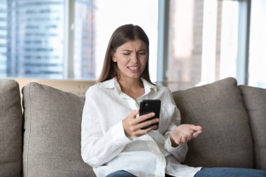 Akıllı telefondan rahatsız olan genç bir kadın, koltukta oturup modern cihaz ekranına bakarken spam aramasından rahatsız oluyor, taburcu oluyor ya da bozuk bir cihaz kullanıyor, SMS 'den kötü haberler alıyor.