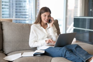 Çekici genç kadın evdeki kanepede dinleniyor, dizüstü bilgisayar kullanıyor, akıllı telefonu tutuyor, uzaktan konuşmanın tadını çıkarıyor ve hoparlörden arkadaşına konuşuyor. Genç nesil ve modern teknoloji kullanımı. Çağrı, iletişim, yardım hattı