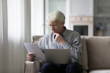 Ciddi derecede endişeli emekli adam evde evrak okuyor, fatura, ödeme faturası, sağlık sigortası anlaşmasını gözden geçiriyor, online uygulamayı laptopta kullanıyor,