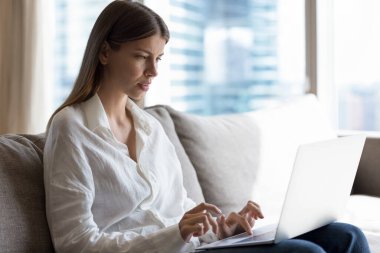 Çalışan ya da ders çalışan, internetten iletişim kuran, sosyal medyada mesajlar paylaşan çekici genç bir kadın modern evdeki kanepede oturuyor. Telework, freelance kablosuz teknoloji ve internet