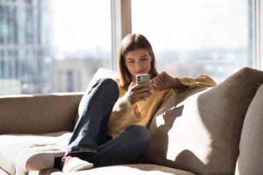 Çekici bir kadın modern akıllı telefonu kullanarak kanepede dinleniyor, internette tembel bir hafta sonu geçiriyor, erkek arkadaşıyla sohbet ediyor, sosyal medya kuryesi aracılığıyla internetten iletişim kuruyor, arama yapıyor, evde sipariş veriyor.