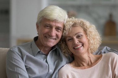 Çekici iyimser yaşlı eşlerin portresi, yakın çekim. Mutlu evlilik, ahenkli ilişkiler, ev sahiplerinin sevecen çifti geniş dişli bir gülümsemeyle modern evde poz veren kameralara bakıyorlar. Aşk