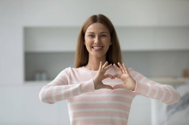 Gülümseyen genç bir kadın kalp işaretini parmaklarıyla gösteriyor. Minnettar poz veriyor. Evde tek başına kameraya bak. Gönüllü bir kadın destek ve şefkat gösteriyor. Aşkın, merhametin ve minnettarlığın sembolü.