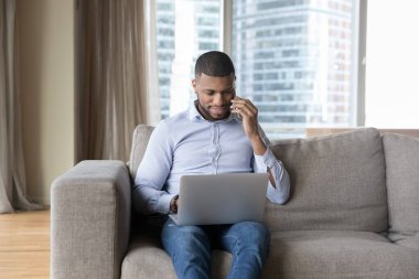 Afrikalı işadamı evden iş yapıyor, koltukta oturuyor cep telefonuyla konuşuyor dizüstü bilgisayarla konuşuyor, müşteriyle görüşüyor, sipariş veriyor, müşteri hizmetleri müdürüyle konuşuyor, modern teknoloji kullanıyor.