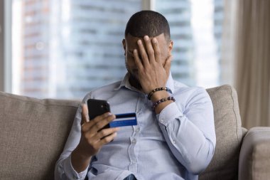 Unsuccessful payment, internet scam, fraud victim. Upset African guy sit on sofa holds card and smartphone, cover face with palm feels shocked, overspending money, lost savings, money stolen from bank clipart