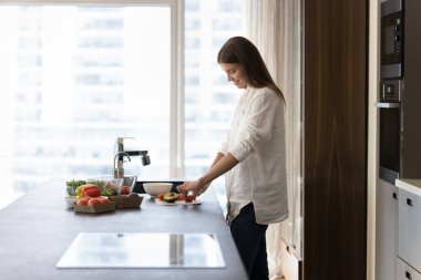 Millennial 30 'ların ev hanımı şık bir mutfakta vejetaryen yemeği pişiriyor. Günlük giysiler içinde genç çekici bir kadın sağlıklı beslenme salatası için sebze keser, evde ev yapımı yemek hazırlar.