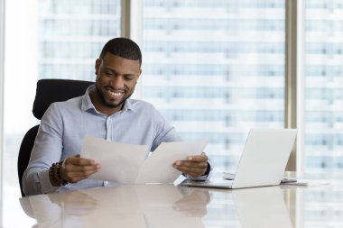 Üzerinde çarşaflar olan Afrikalı iş adamı, ofis masasında oturan evrak raporlarını okuyor, kağıt işleri CEO 'su iyi iş sonucu, iş büyümesi ve satış artışından memnun.