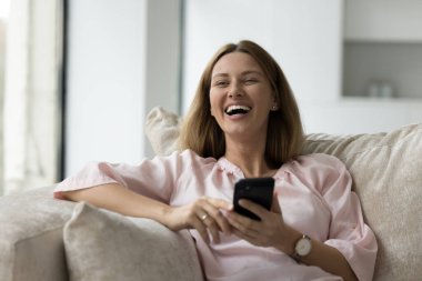 Gülümseyen genç bir kadın evdeki kanepede dinleniyor, gülüyor, internetten alışveriş yapıyor, akıllı telefon kullanıyor, modern cep telefonu kullanan mutlu bir kadın, hoş mesajlar veya mesajlar alıyor, internette sohbet ediyor, keyfine bakıyor ve teknolojinin tadını çıkarıyor.