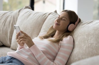 Huzurlu genç bir kadın, radyo kulaklıklarıyla en sevdiği müziği dinliyor, akıllı telefonuyla kanepeye uzanıyor, tasdikler haftasonunu evde modern teknoloji kullanarak geçiriyor. İnternet akışı çevrimiçi hizmetler, dinlenme