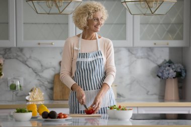 Çekici sarışın orta yaşlı kadın sağlıklı akşam yemeği gülümsemesi için kırmızı biber keser. Ev yapımı vejetaryen yemekleri hazırlar, modern mutfakta tek başına yemek pişirir, ailenin diyetle beslenmesini sağlar.