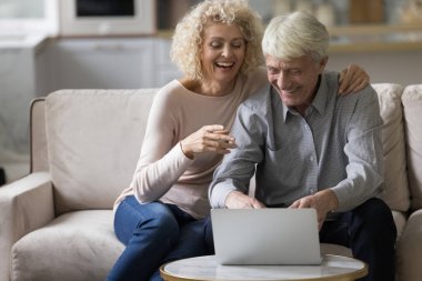 Yaşlı ve çekici bir çift evde oturup kanepede oturup güler, dizüstü bilgisayara bakar, film izler, internetten eğlenceli bir vlog içeriği izler, kablosuz teknoloji ve modern cihazın keyfini çıkarırlar.