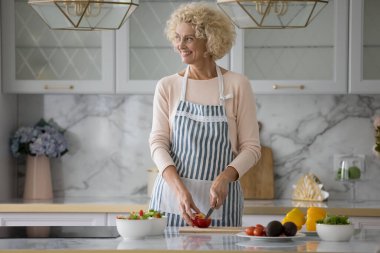 Çekici sarışın orta yaşlı kadın sağlıklı akşam yemeği gülümsemesi için kırmızı biber keser. Ev yapımı vejetaryen yemekleri hazırlar, modern mutfakta tek başına yemek pişirir, ailenin diyetle beslenmesini sağlar.