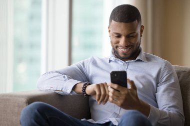 Modern akıllı telefon kullanan yakışıklı Afrikalı adam, mesajlar yolluyor, arkadaşlarıyla mesajlaşıyor, bekarlar için e-randevu servislerini kullanıyor evdeki rahat koltukta dinleniyor. Teknoloji kullanımı, internet, bağlantı