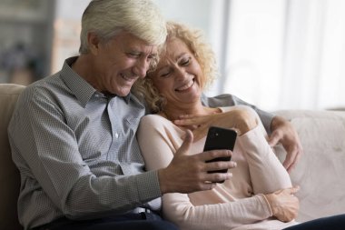 Neşeli olgun yaşlı çift evde cep telefonu kullanıyor, video görüşmesi yapıyor, iyi haberler alıyor, birlikte tatlı online içerik izliyorlar, internet iletişimi için akıllı telefonu paylaşıyorlar, sohbet etmekten zevk alıyorlar.