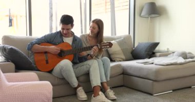Sevgilisini etkilemek için evde akustik gitar çalan mutlu genç bir Latin. Pozitif İspanyol kız müzisyen erkek arkadaşıyla çıkıyor, kanepede oturuyor, müzik dinliyor, konuşuyor.