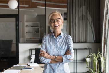 Başarılı orta yaşlı iş kadını, proje lideri ya da akıl hocası kollarını kavuşturmuş, gülüyor, modern işyerinde kameraya poz veriyor, profesyonellik ve pozitiflik yayıyor, ofis portresi yapıyor.