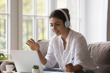 Kablosuz kulaklıklarıyla genç Hintli bir kız öğrencinin bilgisayarda öğretmeniyle video görüşmesi yapması, evdeki oturma odasına notlar yazması, merhaba eli yazması, çevrimiçi bilgisayar eğitimi alması.