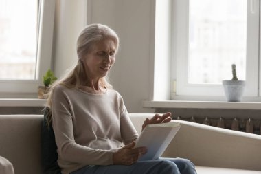 Mutlu yetişkin tablet kullanıcısı kadın dijital cihaz üzerinde sanal uygulama kullanıyor, kitap okuyor, ekrana dokunuyor, boş vakitlerin tadını çıkarıyor, kablosuz iletişimin keyfini çıkarıyor, evde kanepeye oturuyor.