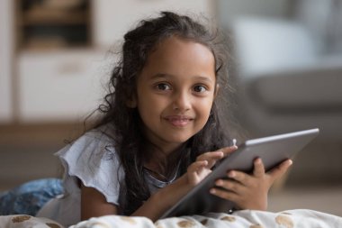 Mutlu tatlı Z jenerasyonu küçük kız tablet bilgisayar tutuyor, kameraya bakıyor, gülümsüyor, yumuşak yastığa sarılıyor, online öğrenme uygulaması kullanıyor, okul ödevi yapıyor, dokunmatik ekrana dokunuyor. Ev resmi