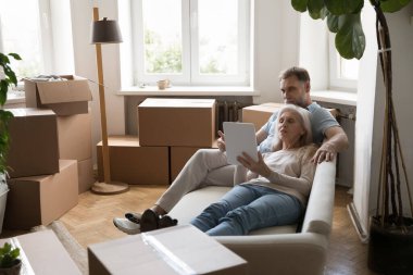 Rahatlamış, gri saçlı birkaç ev sahibi tablet üzerinde grafik uygulama kullanıyor, yeni bir daireye taşındıktan sonra iç tasarım planlıyor, yığılmış kutuların arasında koltukta dinleniyor.