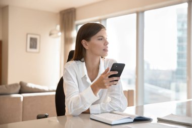 Ciddi bir milenyum iş kadını cep telefonu tutuyor, ofisteki çalışma masasında oturuyor, pencereye bakıyor, düşünüyor, mesaj yazıyor, internette sohbet ediyor, iletişim için cihaz kullanıyor.