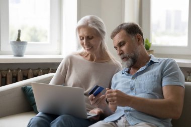Mutlu orta yaşlı koca ve karı koca internetten alışveriş yapıyor, kredi kartıyla online alışveriş yapıyor, dizüstü bilgisayardaki banka ekommerce uygulamasını kullanıyor, evdeki kanepede dinleniyor.