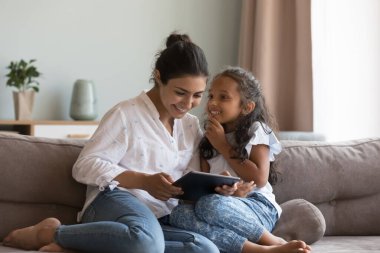 Mutlu Hintli kız çocuğu annesiyle konuşuyor, dijital tablet kullanıyor, koltukta oturuyor, gülümsüyor, gülüyor. Anne ve çocuk eğleniyor, dijital aletlerle oyun oynuyorlar.