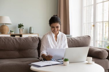 Laptop 'ta kağıt okuyan mutlu Hintli çalışan kadın, iyi haberlere gülümsüyor, satış raporları, iş sonuçları, sözleşme, anlaşma koşulları, dizüstü bilgisayarda koltukta oturmak, evde çalışmak.