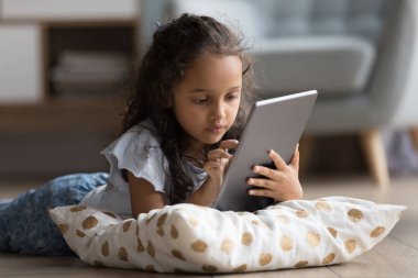 Odaklanmış Z geneli okul çocuğu yumuşak yastıkla ısıtma zemininde dinleniyor, dijital tablet bilgisayarda öğrenme uygulaması kullanıyor, kitap okuyor, ödev yapıyor, internette geziniyor, sosyal medyada geziniyor.