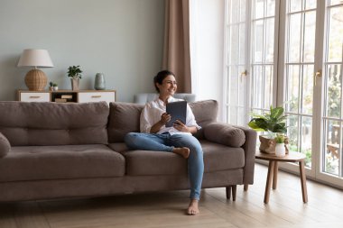 Mutlu Hintli alet kullanıcısı kadın dijital ped tutuyor, kanepede dinleniyor, iyi düşüncelerle pencereye bakıyor, gülümsüyor, tablet üzerinde kitap düşünüyor, keyfine bakıyor, evde rahat ediyor.