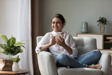 Neşeli, genç, Hintli bir kadın hücrede oturuyor, koltuğunda oturuyor, başka yere bakıyor, gülümsüyor, gülüyor, sohbet ediyor, daktilo ediyor, internette uygulama yazıyor, kısa mesaj üzerinden düşünüyor.