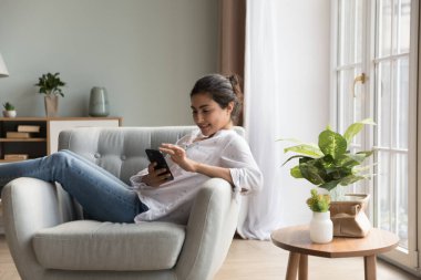 Neşeli, rahat, cep telefonu kullanan genç Hintli kadın, modern evdeki rahat koltukta dinleniyor, internet ekommerce uygulaması ile alışveriş yapıyor, ekrana dokunuyor, gülümsüyor.