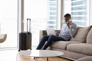 Etkili bir Afrikalı gezgin iş seyahatinde otel odasında iş iletişimi için aletler kullanıyor, dizüstü bilgisayar kullanıyor, bavul başında cep telefonuyla konuşuyor.