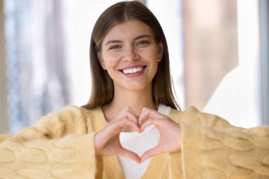 Neşeli, genç ve güzel bir kadın, kalbini göğsünde tutuyor, el hareketleriyle aşk, romantizm, sevgililer günü, romantik duygular paylaşıyor, dişlek bir gülümsemeyle kameraya bakıyor.