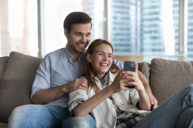 Güzel, mutlu, milenyum aşığı bir çift kanepeye uzanıp cep telefonu ekranına bakıyor komik videolar izliyor, mobil video uygulaması ile ailesiyle video konferans görüşmesinin keyfini çıkarıyor, modern teknoloji kullanarak boş vakitlerini geçiriyor.