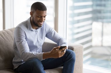 Neşeli çekici genç siyahi adam evde internet iletişiminden zevk alıyor, cep telefonu kullanıyor, online uygulama kullanıyor, şehirdeki dairede kanepede oturuyor, ekrana bakıyor, gülümsüyor.