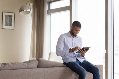 Mutlu yakışıklı Afrikalı adam tablet ekrana dokunuyor, kanepeye yaslanıyor, dijital cihazlarda online uygulama kullanıyor, gülümsüyor, gülüyor, internet iletişiminden keyif alıyor.