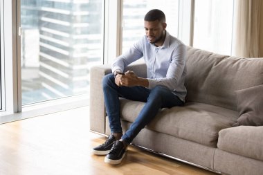 Yakışıklı, genç Afrikalı, akıllı telefonuyla daktilo eden, evdeki koltukta oturan, cihaz üzerinde başvuru yapan, kentsel apartman dairesindeki iş görevleri üzerinde çalışan.